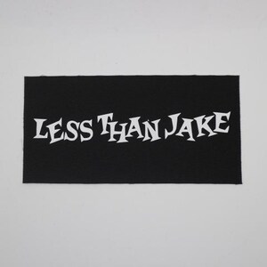 May include: A black rectangular patch with the white text "LESS THAN JAKE" in a stylized font. The patch is likely made of fabric and is set against a white background.