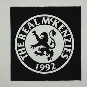 May include: A square black fabric patch featuring a white circular emblem. The emblem displays a central rampant lion silhouette surrounded by the text THE REAL MCKENZIES and the year 1992 printed in a bold, white, sans-serif typeface.