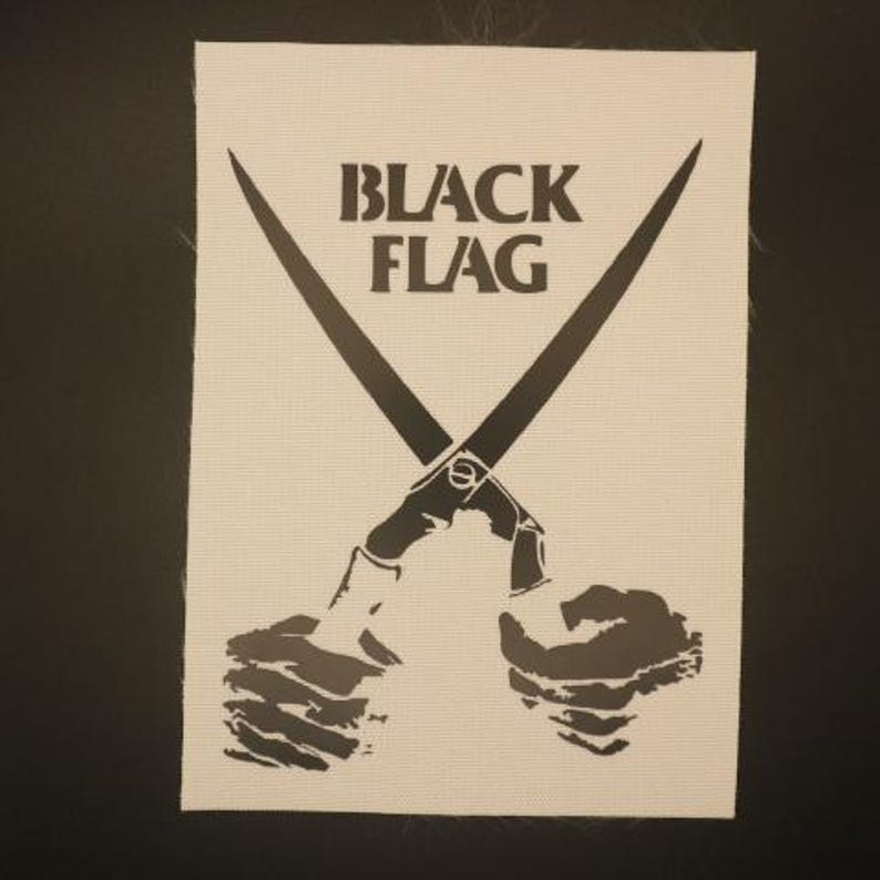 May include: A rectangular fabric patch featuring a cream-colored background with a black graphic print. The design displays two hands holding a pair of crossed scissors, with the text "BLACK FLAG" printed in bold, black capital letters above.