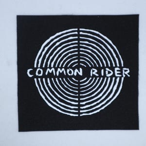 May include: A square black fabric patch featuring a white graphic design of concentric circles centered behind the text COMMON RIDER printed in a bold, sans-serif font that horizontally bisects the circular pattern across the middle.