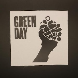 May include: A square, off-white patch featuring the words "GREEN DAY" in bold, black capital letters above a black silhouette of a hand holding a grenade. The background is a textured, light-colored fabric.