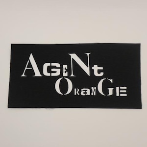 May include: A black rectangular patch with the words "AGENT ORANGE" in white, stencil-style lettering. The patch has a textured surface and is set against a plain white background.