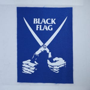 May include: A rectangular blue fabric patch featuring a white graphic print of two hands holding a pair of crossed scissors. The text "BLACK FLAG" is printed in white capital letters above the scissors. The patch has a visible woven texture.