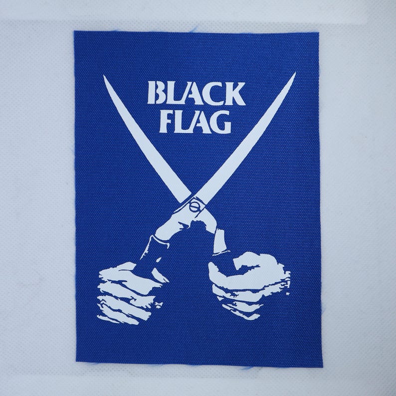 May include: A rectangular blue fabric patch featuring a white graphic print of two hands holding a pair of crossed scissors. The text "BLACK FLAG" is printed in white capital letters above the scissors. The patch has a visible woven texture.