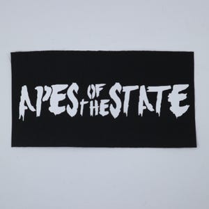 May include: Black rectangular patch with the white text "APES OF THE STATE". The text has a distressed, dripping font style. The patch is made of a textured fabric.