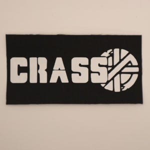 May include: A rectangular black fabric patch featuring the white printed text CRASS in a bold, blocky font, followed by a circular white graphic symbol consisting of a cross shape enclosed within a circle, all set against a plain background.