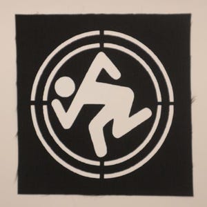 May include: A square black fabric patch featuring a white screen-printed graphic of a stylized human figure in a running or crouching pose, centered within a circular target-like frame with segmented lines.