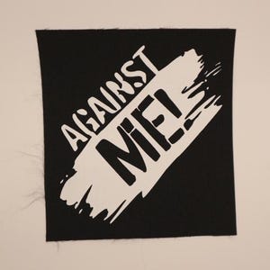 May include: Square black patch with the words "AGAINST MIE!" in white, stylized text. The text is overlaid on a white brushstroke design. The patch has a raw edge.