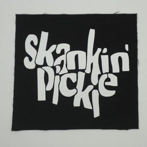 May include: A black square patch with the words "Skankin' Pickle" in large, white, stylized lettering. The text is centered on the patch, which has a slightly rough, textured appearance.