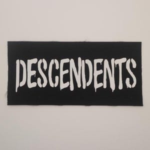 May include: A black rectangular patch with the word "DESCENDENTS" in white, stencil-style lettering. The letters have a dripping effect. The patch is likely for clothing or accessories.