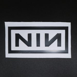 May include: A white rectangular patch with a black border and the letters "NIN" in black. The patch is on a black textured surface.