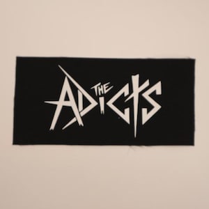 May include: Black rectangular patch with the white text "THE ADICTS". The text is stylized with sharp angles and a unique font. The patch is likely made of fabric and is suitable for sewing or ironing onto clothing or accessories.