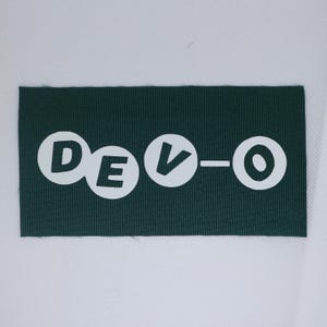May include: A dark green rectangular patch featuring the word "DEVO-O" in white, stylised lettering. The letters are in white circles, with a dash between the "V" and the "O". The patch is made of a textured fabric.