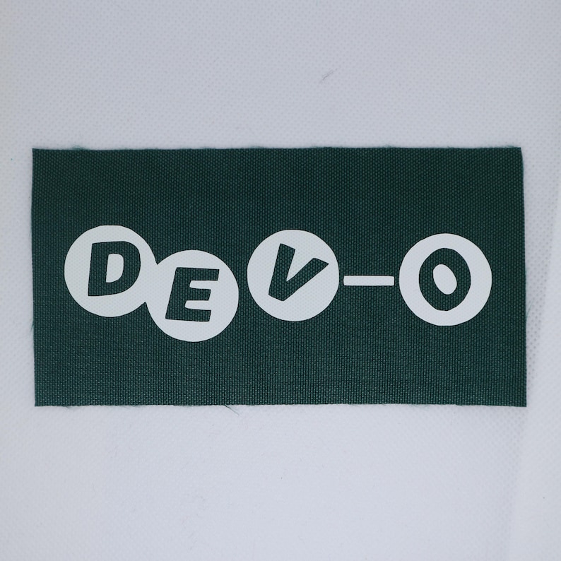 May include: A dark green rectangular patch featuring the word "DEVO-O" in white, stylised lettering. The letters are in white circles, with a dash between the "V" and the "O". The patch is made of a textured fabric.