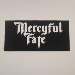 May include: Black rectangular patch with the words "Mercyful Fate" in white gothic lettering. The patch has a textured surface and is likely made of fabric.