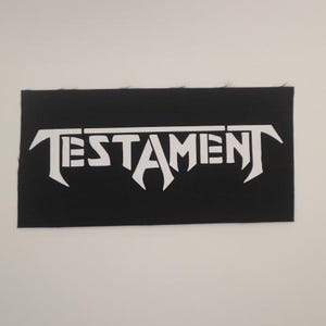 May include: White "TESTAMENT" logo patch on a black fabric background.