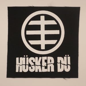 May include: Black square patch with a white graphic design. The design features a circle with a cross-like shape inside and the words "HÜSKER DÜ" in white block letters below the graphic. The patch has a textured surface.