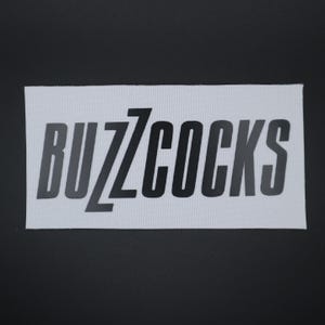 Op de afbeelding: Een rechthoekige witte patch met de tekst "BUZZCOCKS" in zwarte, gestileerde letters. De patch is geplaatst tegen een donkergrijze achtergrond. De stof heeft een subtiele, getextureerde uitstraling.