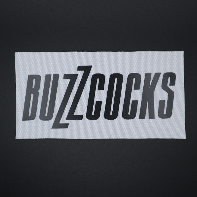 Op de afbeelding: Een rechthoekige witte patch met de tekst "BUZZCOCKS" in zwarte, gestileerde letters. De patch is geplaatst tegen een donkergrijze achtergrond. De stof heeft een subtiele, getextureerde uitstraling.