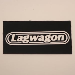 May include: Black rectangular patch with the white text "Lagwagon" inside a white oval border. The patch is made of a textured fabric and is centered on a light background.