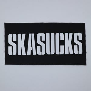 May include: Black rectangular patch with the white text "SKASUCKS" in a bold, sans-serif font. The text is centered on the patch. The patch has a textured appearance.
