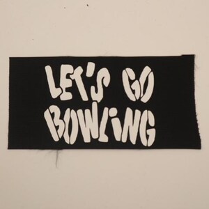 May include: A black rectangular patch with the white text "LET'S GO BOWLING". The text is in a bold, stylized font. The patch has a slightly frayed edge and is set against a neutral background.