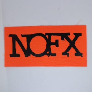Peut inclure: Un &eacute;cusson rectangulaire orange avec le texte noir "NOFX". Les lettres sont en gras et l&eacute;g&egrave;rement us&eacute;es. L'&eacute;cusson est probablement en tissu et se d&eacute;tache sur un fond blanc.