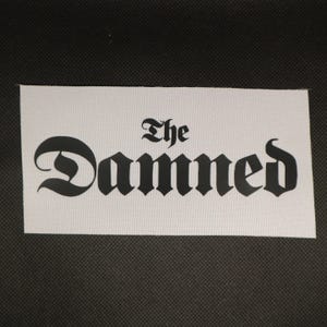 May include: A white rectangular patch with the words "The Damned" printed in black gothic lettering. The patch has a textured surface and is set against a dark background.