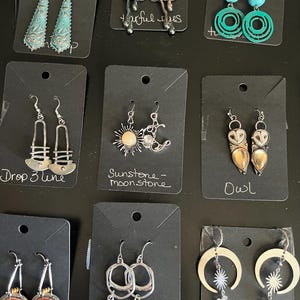 May include: A collection of handcrafted earrings displayed on black cards. Styles include turquoise, sunstone, moonstone, owl, and silver drop designs. Some earrings have text labels like "Drop 3 line" and "Sunstone - moonstone".
