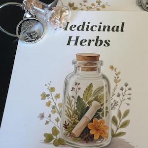 May include: A clear glass bottle-shaped keychain with a cork stopper, filled with dried herbs, next to a card that says "Medicinal Herbs". The card features a watercolor illustration of a glass jar with herbs and a mortar and pestle.