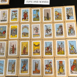 May include: A collection of tarot cards with colorful illustrations and a beige border. The cards are arranged on a black surface. The text "CUPS AND WANDS" is visible at the top. Each card features unique artwork.