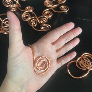 May include: Copper wire art pieces in various swirling shapes. One piece is held in a hand, showcasing a large spiral. Other pieces are scattered on a dark surface, including a series of connected swirls and individual loops. The copper has a warm, reddish-brown hue.