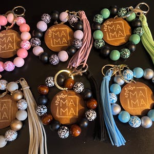 May include: A collection of beaded keychain bracelets in various colors, including pink, black, and blue. Each bracelet features a wooden tag with the word "MA MA" and a tassel. Some beads have animal print patterns.