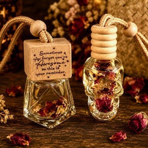 May include: Two hanging glass car air freshener bottles with dried flower petals inside. One bottle is cube-shaped with a wooden cap engraved with the text: Sometimes you forget you're Awesome so this is your reminder. The other is skull-shaped.