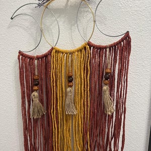 May include: A macrame wall hanging with three crescent moon shapes, featuring tassels and wooden beads in shades of brown, red, and yellow.