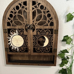 May include: A wooden cabinet with an arched top and two doors. The doors feature intricate cut-out designs. A sun and moon are affixed to the doors. The cabinet has a small shelf at the bottom and is stained a dark brown color.