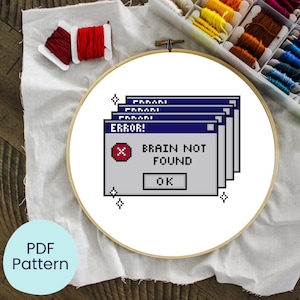 Peut inclure: Un cercle à broder avec un motif de point de croix humoristique. Le motif représente un message d'erreur informatique pixélisé : "ERROR! BRAIN NOT FOUND" avec un bouton "OK". Du fil à broder et une étiquette de patron PDF entourent le cercle.