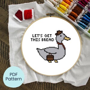 Può includere: Un telaio per punto croce con un disegno di oca pixelata, con cappello e valigetta, con la scritta "LET'S GET THIS BREAD". Il telaio è su tessuto bianco, con rocchetti di filo rosso e una scatola di filo da ricamo colorato.