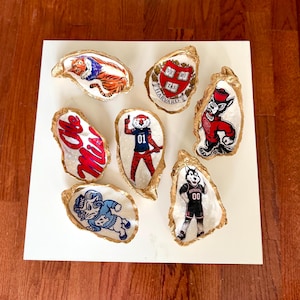 May include: A collection of six decorative oyster shells with gold-painted edges, each featuring hand-painted collegiate imagery including the Harvard crest, Ole Miss text, and various university mascots like a tiger, ram, and husky figures.