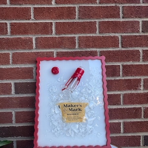 May include: A framed artwork featuring a Maker's Mark whiskey bottle and ice cubes. The frame is red with a scalloped edge. The bottle and ice are set against a white background. The Maker's Mark label is visible.