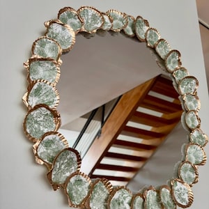 May include: Round mirror with a decorative border of seashells. The seashells are white with a green floral pattern and are edged with a metallic gold color. The mirror is hanging on a white wall.