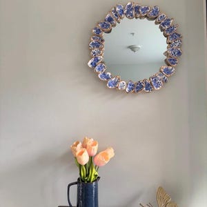 May include: Round mirror with a gold-colored frame and a border of blue and white porcelain pieces. A navy blue pitcher with peach-colored tulips and a gold butterfly decoration are on a wooden surface below the mirror.