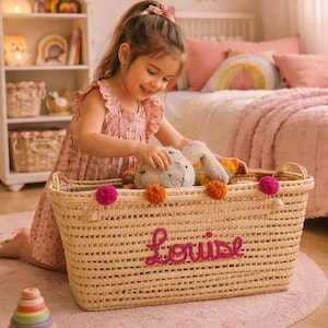 Peut inclure: Scène de chambre d'enfant avec un panier en osier brodé du nom "Louise" en rose. Le panier est rempli de jouets et décoré de pompons roses et oranges. Une enfant en robe rose joue avec les jouets.