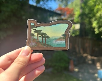 Könnte beinhalten: Ein Aufkleber in Form des Bundesstaates Oregon, der eine Küstenlandschaft mit Bäumen, Meereswellen und einem Leuchtturm zeigt. Der Aufkleber hat einen braunen Rand und ein Design im Aquarellstil.