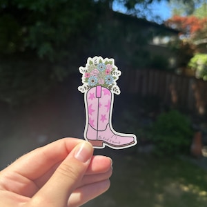 May include: A sticker featuring a pink cowboy boot with star accents, filled with a bouquet of flowers in shades of pink, blue, and green. The text "Be kind 2 yourself" is written on the boot. The sticker is held in a hand.