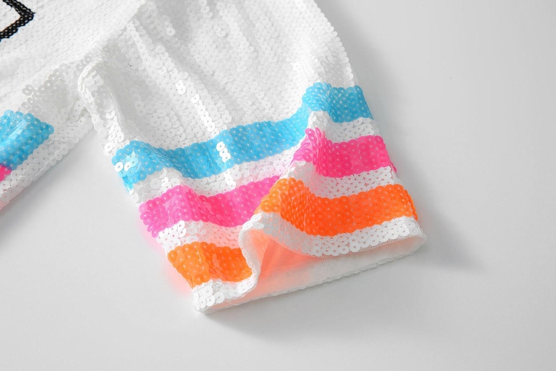 May include: White sequined fabric with colorful horizontal stripes in blue, pink, and orange. The fabric appears to be part of a garment, possibly a sleeve or trim, with a bright and playful design.