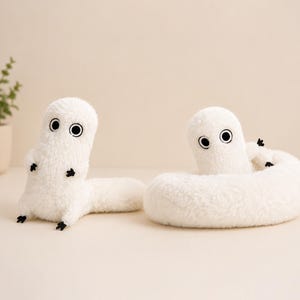 May include: Two white plush toys shaped like worms or caterpillars with black eyes and tiny black feet. One is upright, the other coiled. The toys are on a light beige surface.