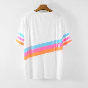 May include: White sequin t-shirt with a colorful diagonal stripe across the front. The stripe includes bands of orange, pink, blue, and orange sequins. The sleeves have matching stripes of orange, pink, and blue sequins.