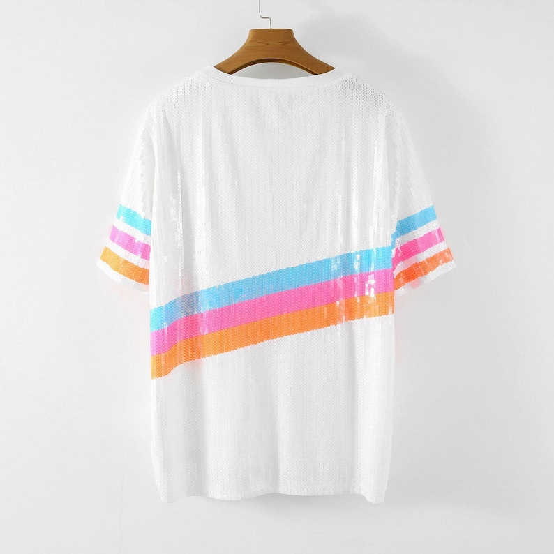 May include: White sequin t-shirt with a colorful diagonal stripe across the front. The stripe includes bands of orange, pink, blue, and orange sequins. The sleeves have matching stripes of orange, pink, and blue sequins.