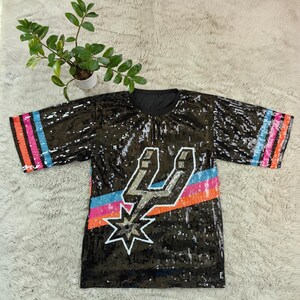 May include: Black sequined t-shirt with a colorful design. The shirt features a silver lightning bolt and star design, with a blue, orange, and pink stripe. The sleeves have matching stripes.